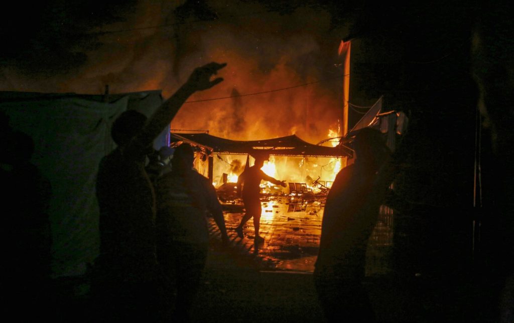 People silhouetted against a large fire at night, with flames engulfing structures in the background. Figures are raising their hands as others move towards the blaze. The scene is chaotic, with heavy smoke in the air. This image is associated with the "Al-Aqsa Hospital attack."