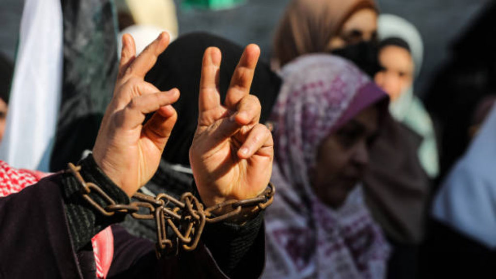 A Palestinian woman with chained hands raises the peace symbol during a protest, symbolizing the ongoing struggles of Palestinian women fighting for freedom amidst the violence of war.